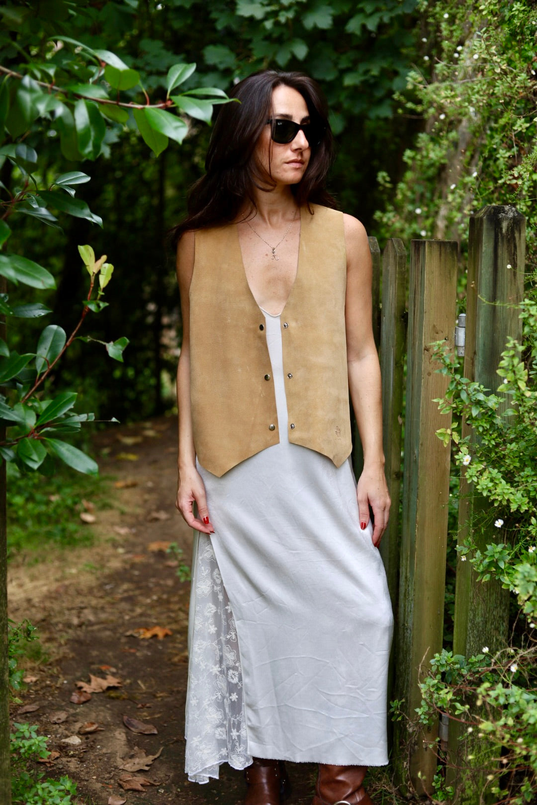 A woman stands outdoors on a wooded path wearing a tan suede vest over a white top and a long light gray skirt, with brown boots and dark sunglasses.