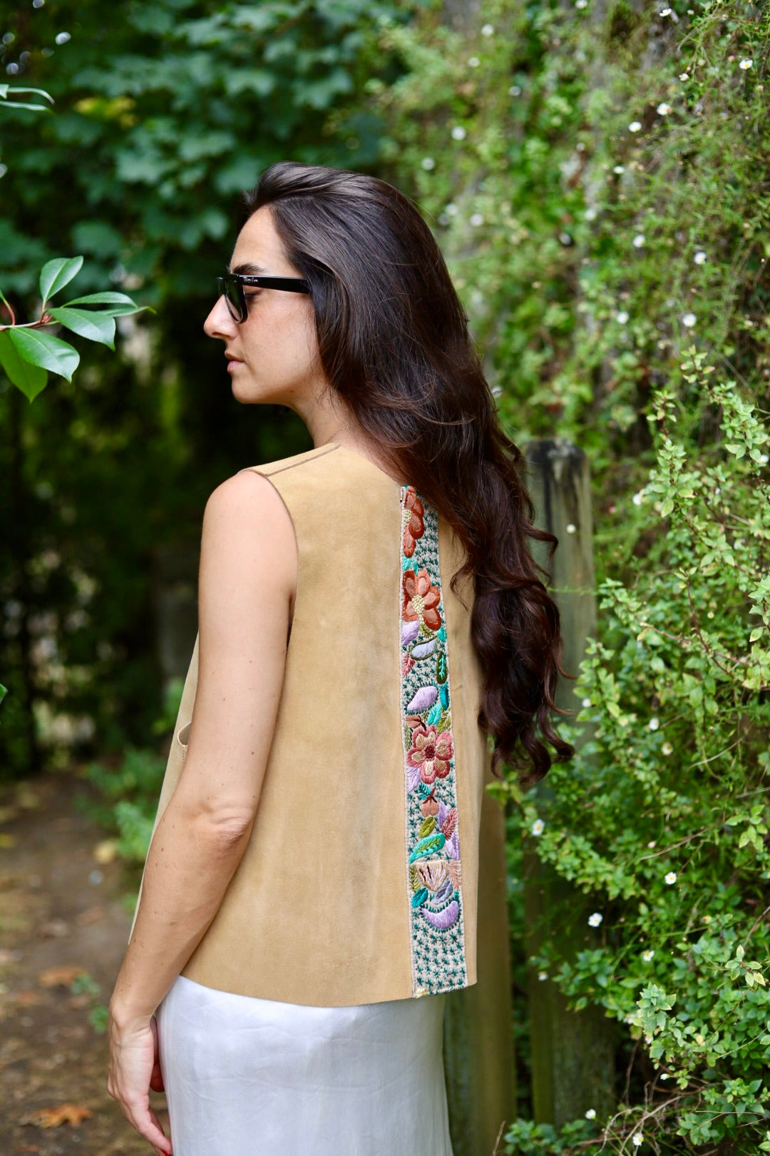 A woman wearing a sleeveless tan vest with a colorful floral embroidered vertical panel on the back, photographed outdoors against a lush green garden background.