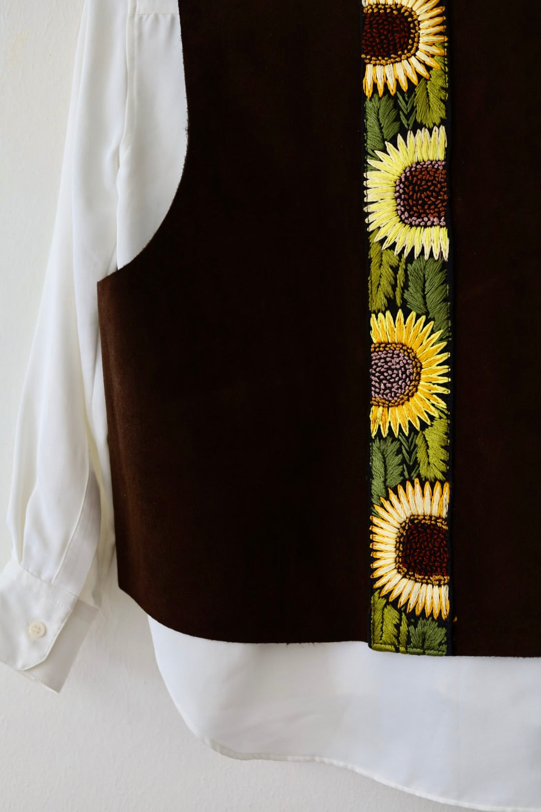 A close-up of a dark brown vest layered over a white button-up shirt, featuring a vertical embroidered sunflower pattern in yellow, green, and brown.
