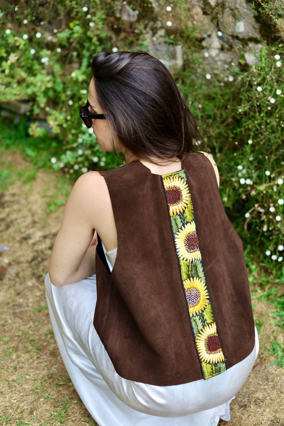A woman wearing a brown suede vest with a vertical sunflower print panel on the back, photographed outdoors in a garden setting.