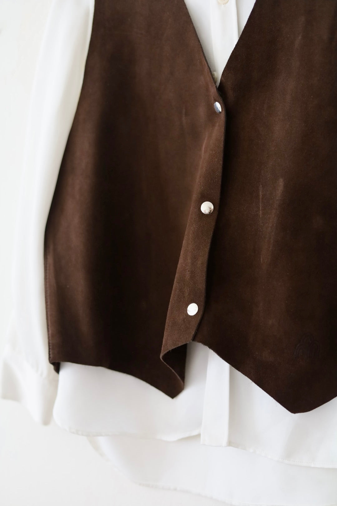 A close-up of a dark brown suede vest layered over a white button-up shirt, featuring three silver snap buttons.
