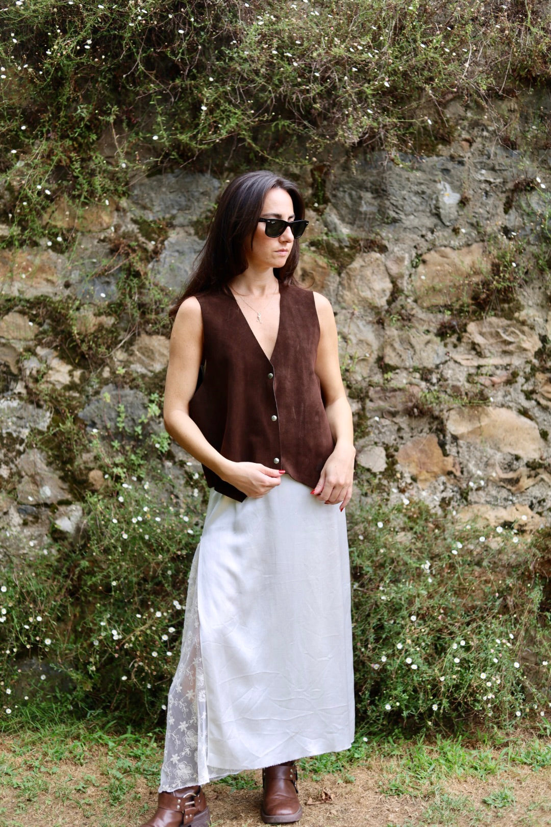 A woman wearing a sleeveless brown vest and a long light gray skirt stands outdoors in front of a stone wall with greenery.