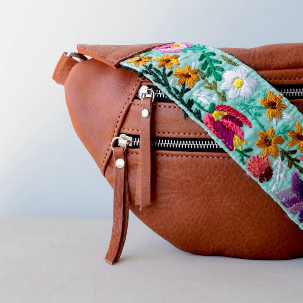 A brown leather sling bag with a caramel color and a floral-embroidered strap, featuring two front external pockets.