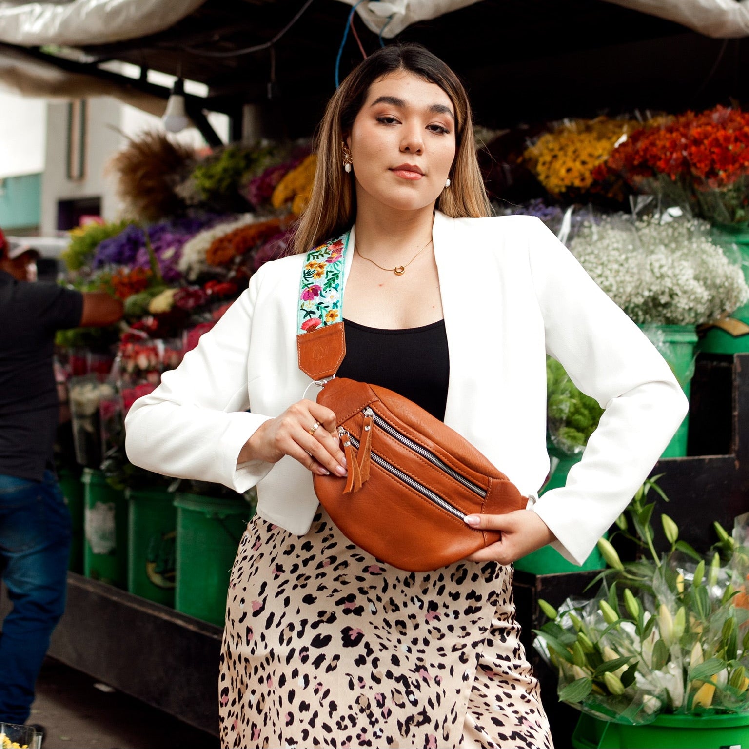 Woman holding a brown leather bag in front of a market stall with flowers and produce.