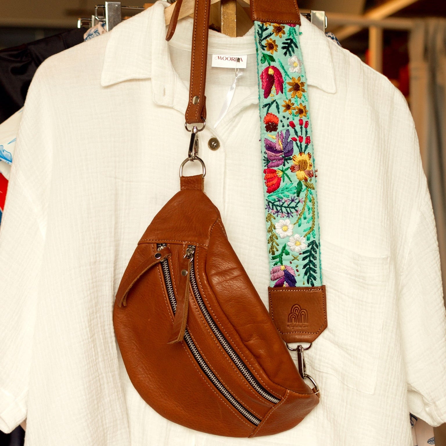 Brown leather backpack with a colorful floral strap on a hanger against a white shirt background.