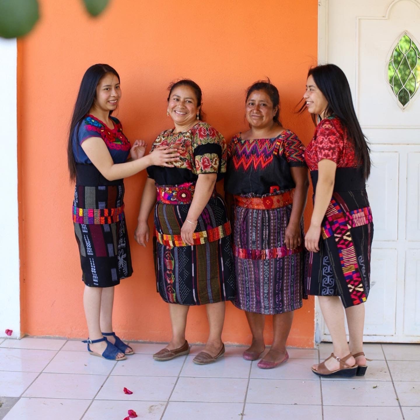 Four women in traditional attire standing in front of an orange wall.