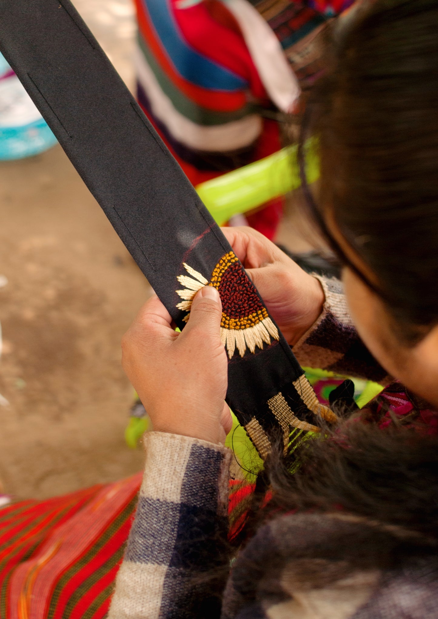 A close-up of a person's hands holding a black woven textile, possibly a bag strap or decorative trim, and applying a white and yellow sunflower bead or embroidery design. The person is wearing a gray and navy plaid sweater, and a colorful red, green, and blue patterned textile is visible in the background.