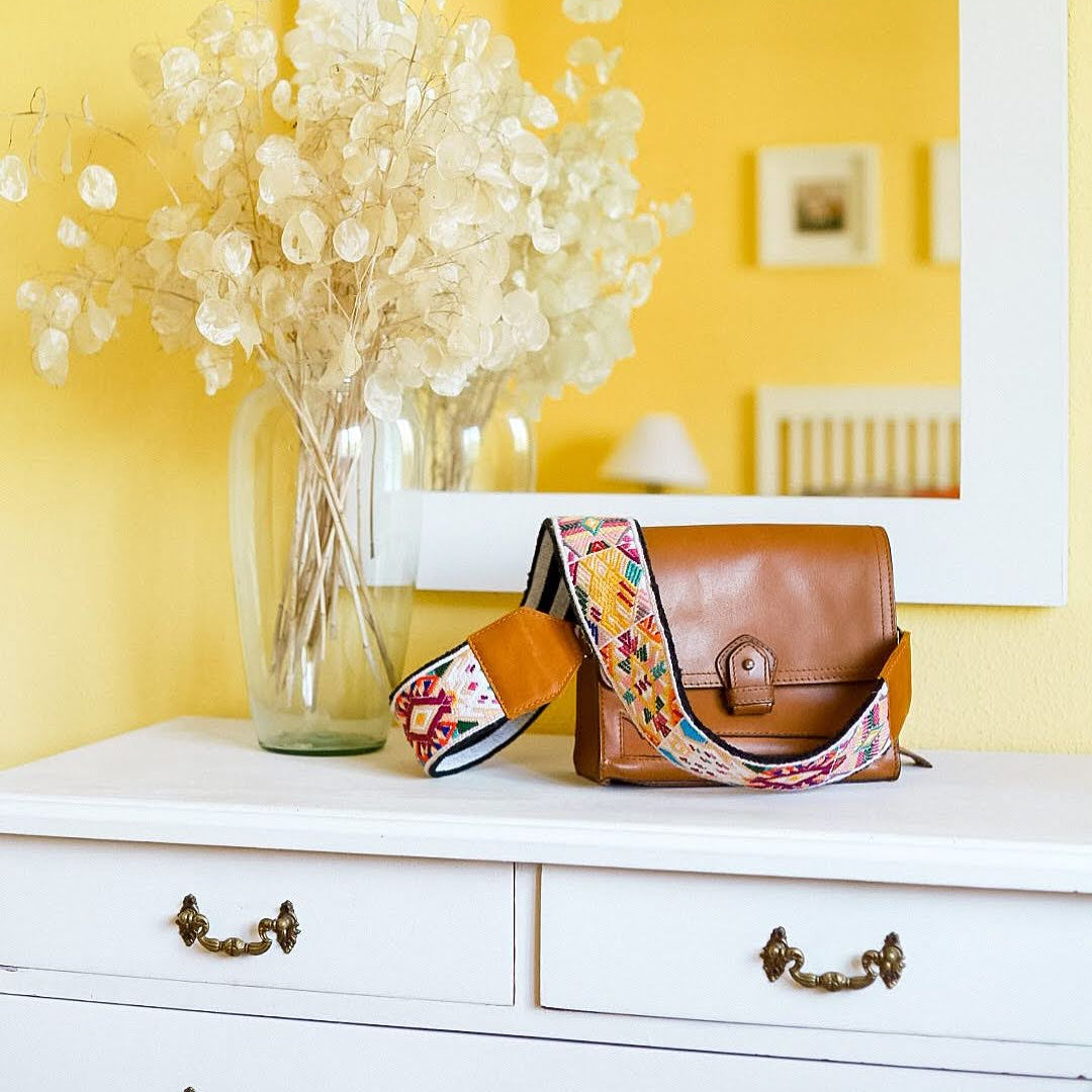 Brown leather bag with colorful strap on a white dresser against a yellow wall.