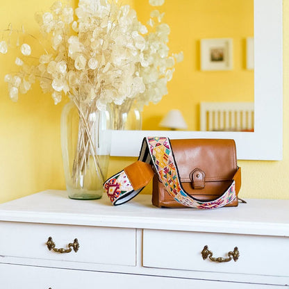 Brown leather bag with colorful strap on a white dresser against a yellow wall.