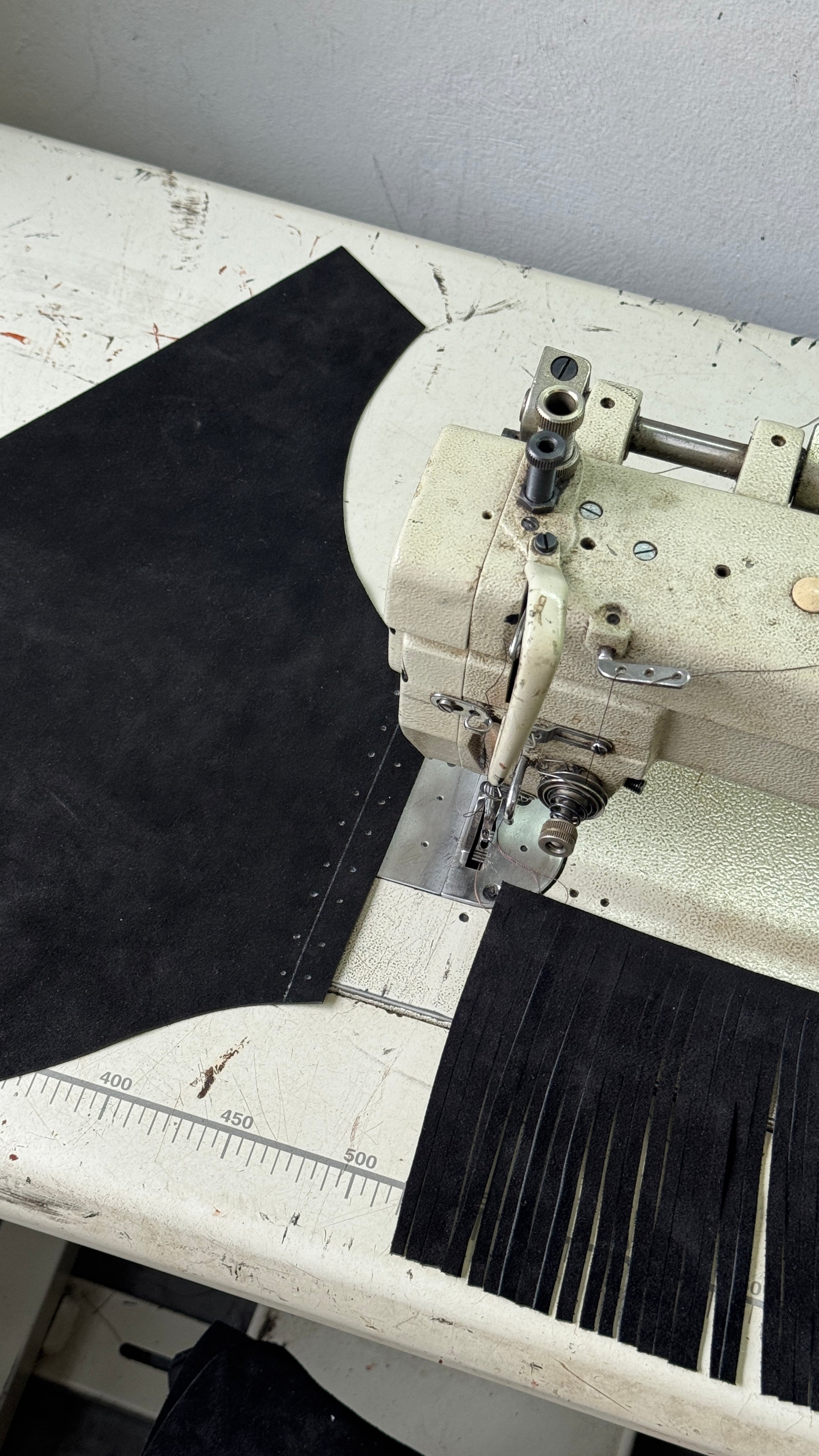 A close-up of a vintage cream-colored industrial sewing machine on a worn white worktable, with a large black fabric piece and a strip of cut black fringe material beside it.