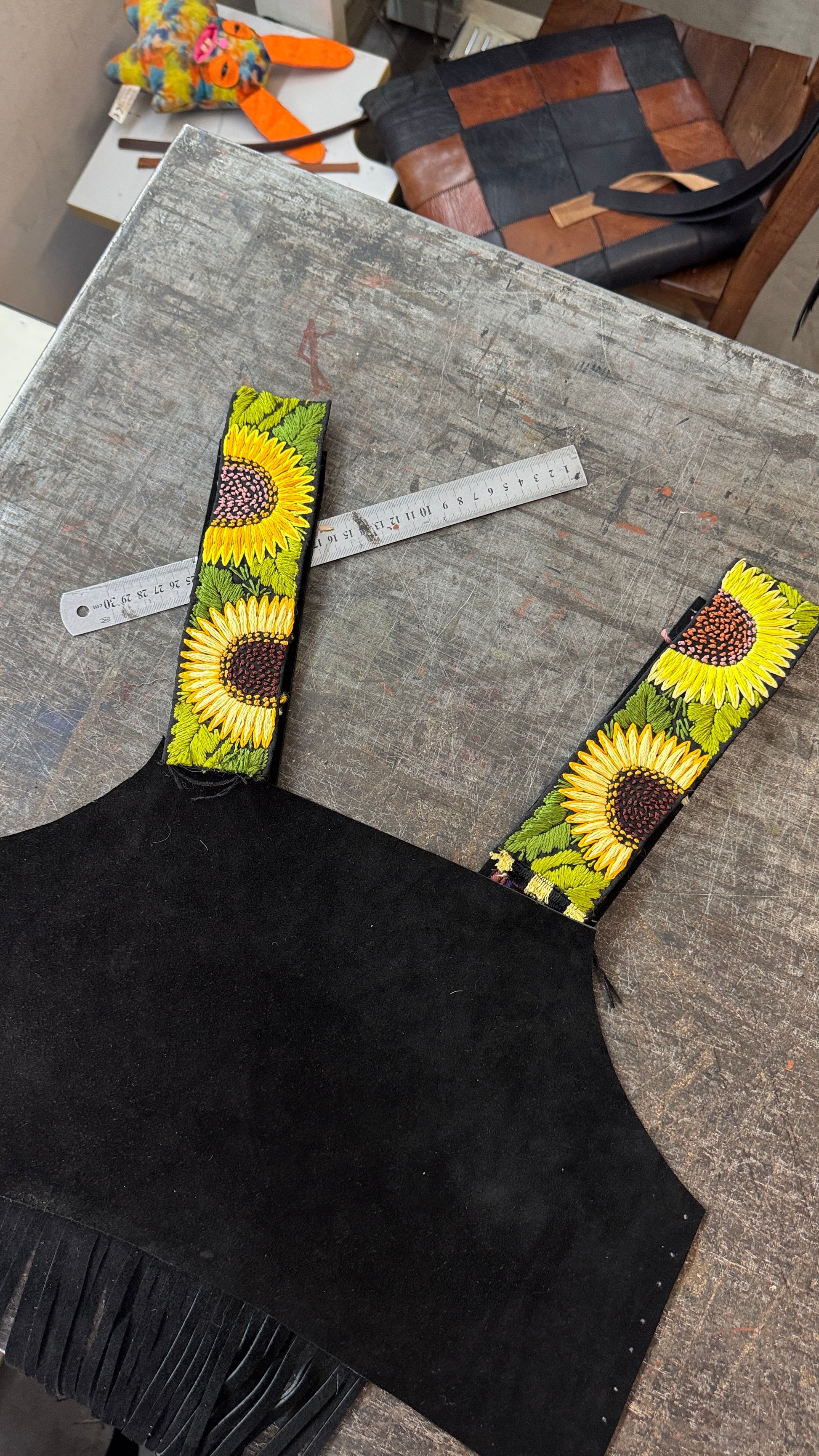 A black suede-like vest or top with wide shoulder straps featuring a bright sunflower print, laid flat on a gray wooden table with a metal ruler nearby.