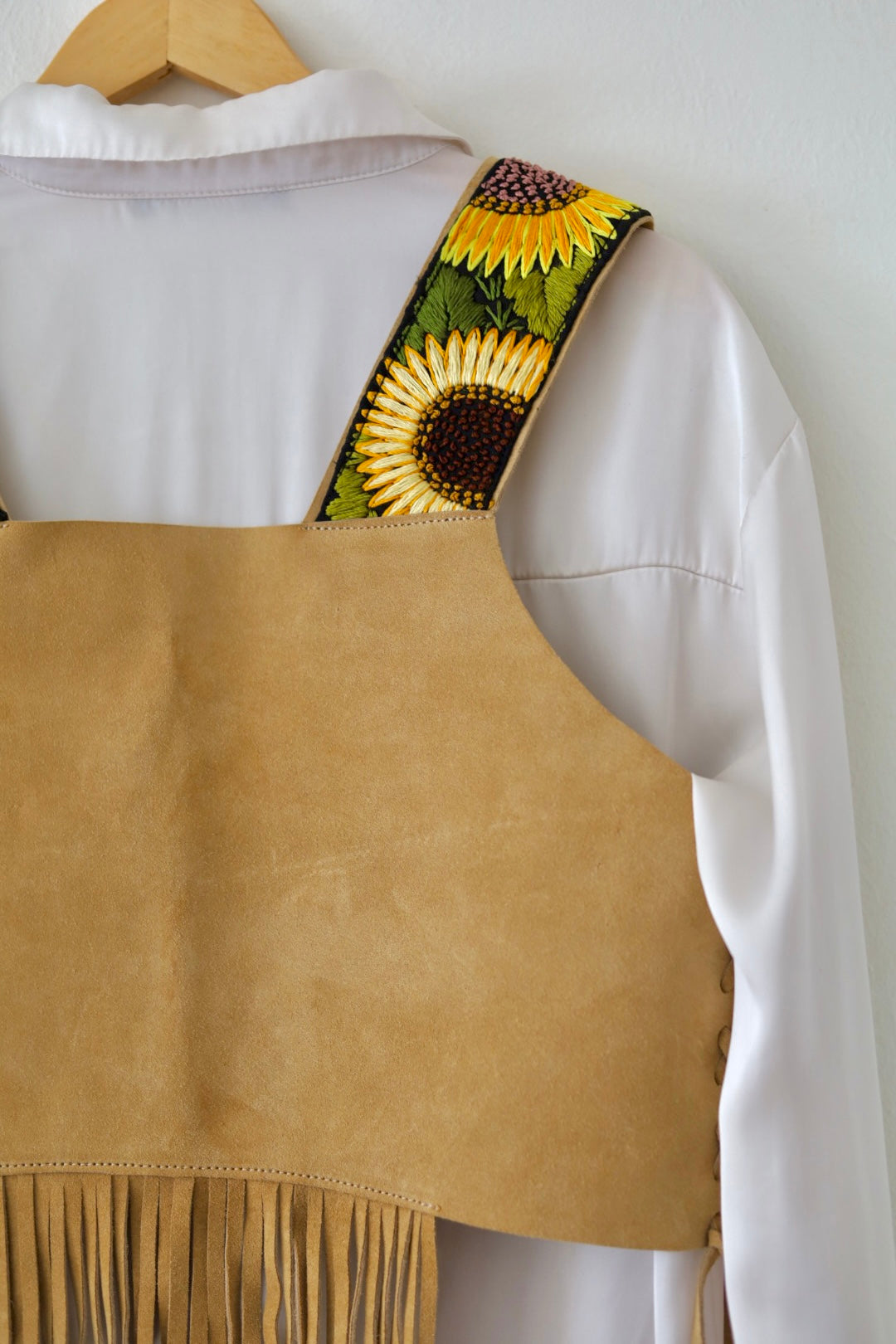 A tan suede fringed vest or apron-style garment hanging on a wooden hanger over a white shirt. The shoulder strap features a printed sunflower pattern with green leaves and dark centers.