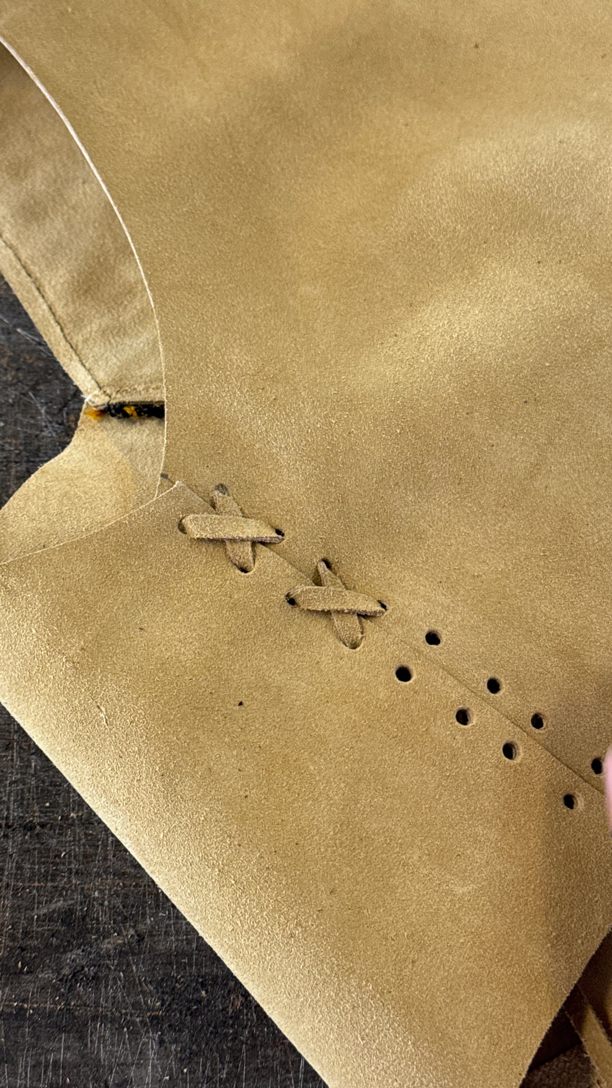 Close-up of a tan suede or leather garment piece with decorative stitching and eyelet holes, laid on a dark textured surface.