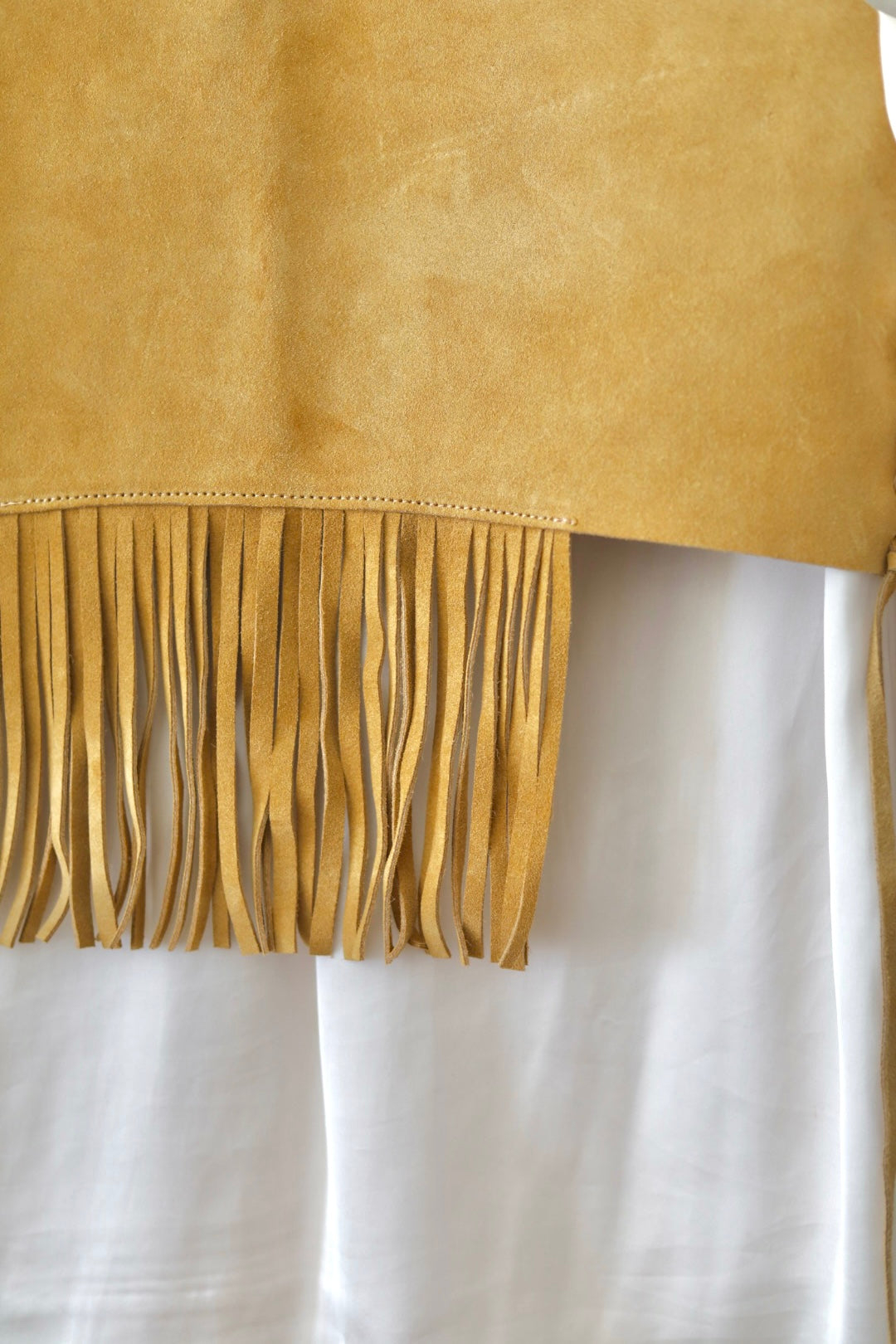 A close-up of a mustard yellow suede fringe garment, likely a shawl or wrap, draped over a white fabric background.