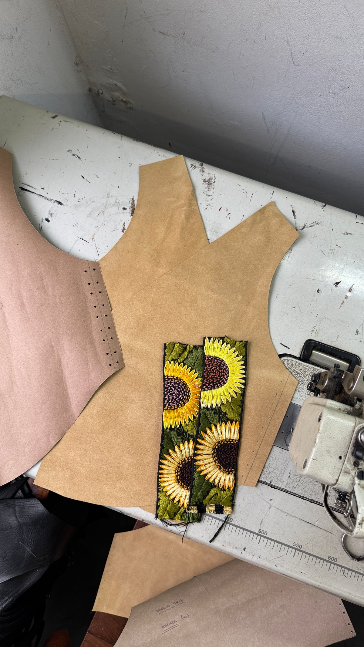 A pattern cut for a sleeveless garment laid on a white worktable, alongside a matching fabric swatch featuring a sunflower print. A sewing machine is partially visible on the right.