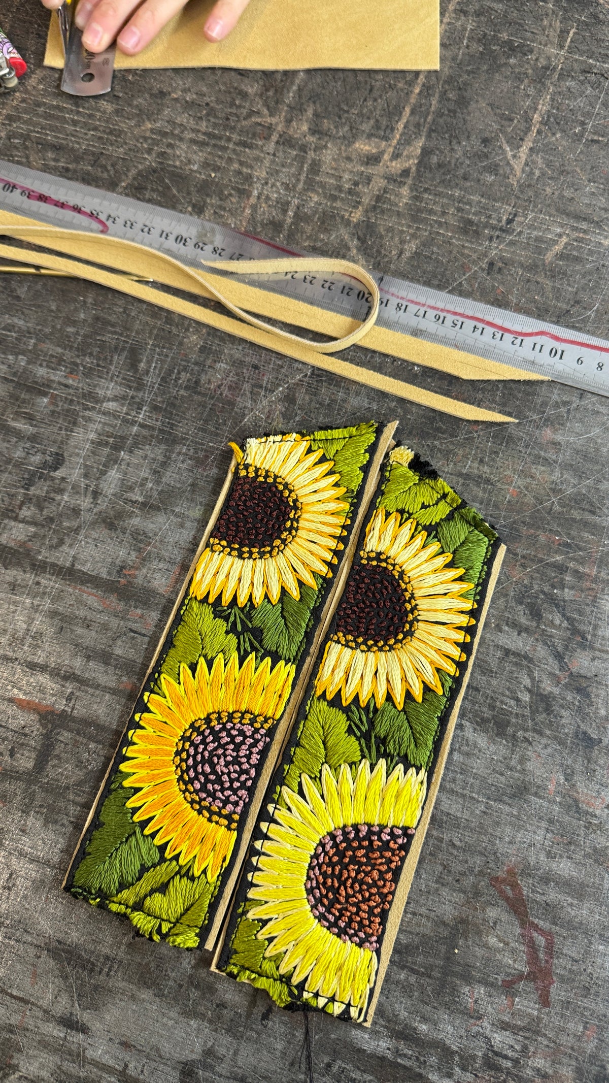 Two rectangular leather pouches decorated with a vibrant yellow and green sunflower print, laid flat on a dark wood surface next to a metal ruler and leather strips.