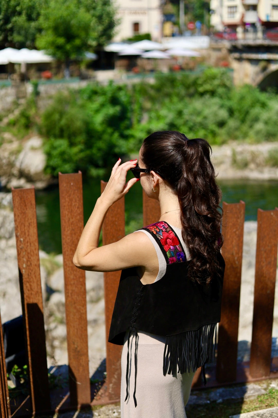 A woman with long dark hair in a ponytail stands outdoors by a railing, wearing a sleeveless black top with colorful embroidered shoulder details and fringe, paired with a light beige skirt. The background shows green foliage, water, and a distant urban setting.