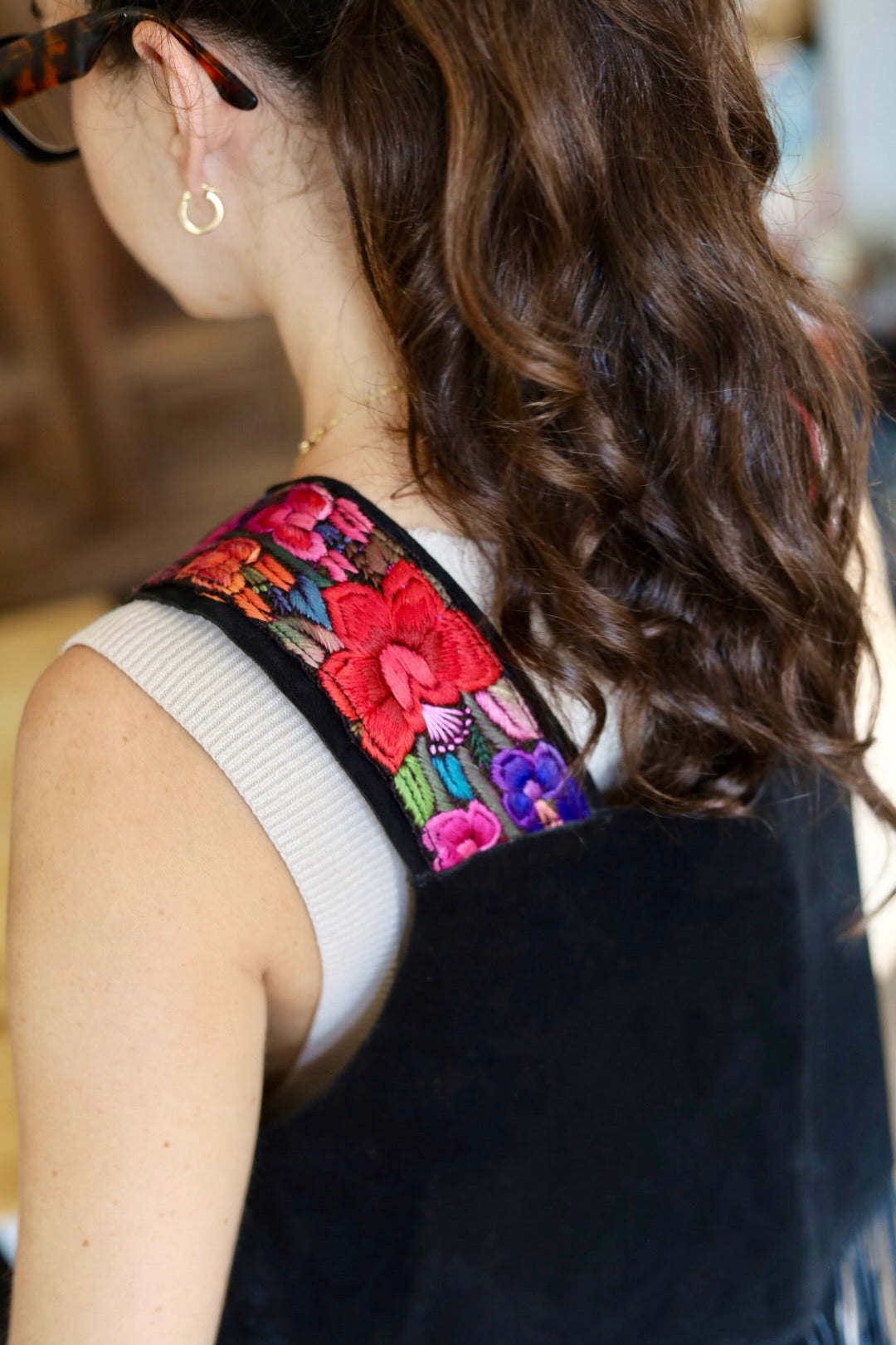 Close-up back view of a woman wearing a black sleeveless top with colorful floral embroidery across the shoulder, over a white ribbed undershirt. She has long wavy brown hair and wears a small gold hoop earring.