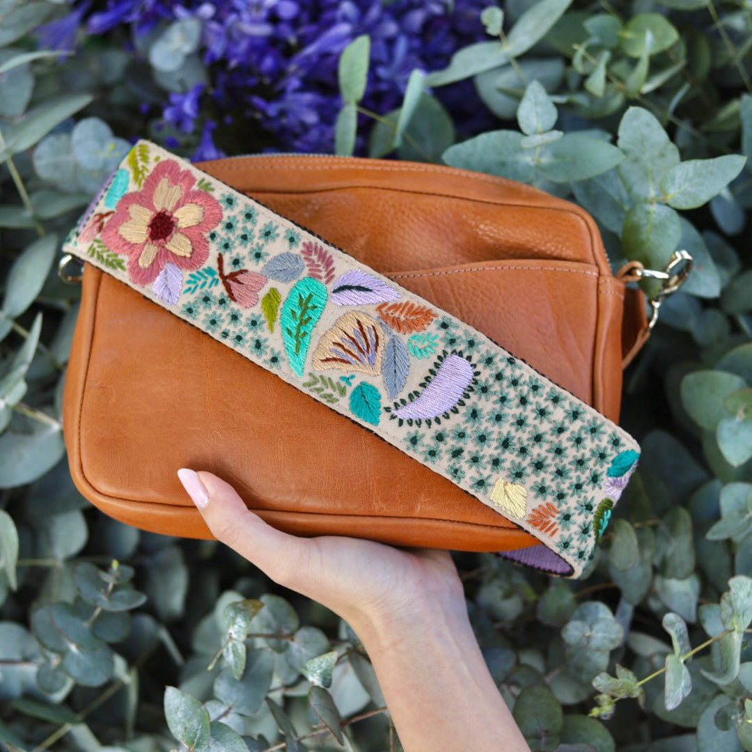 Brown leather handbag with a floral strap held by a hand against a green leafy background