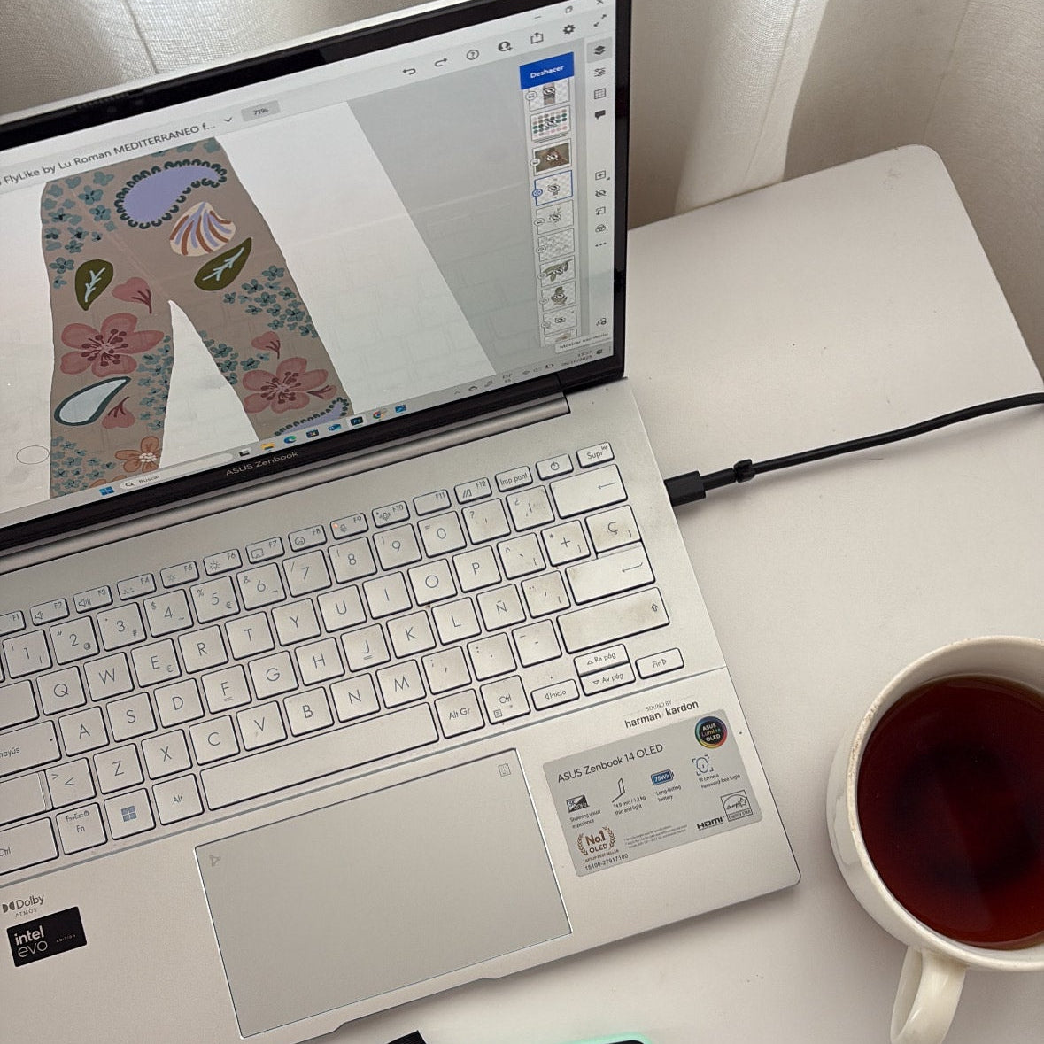 Laptop with graphic design software on screen, stylus, and mug of tea on a white surface.
