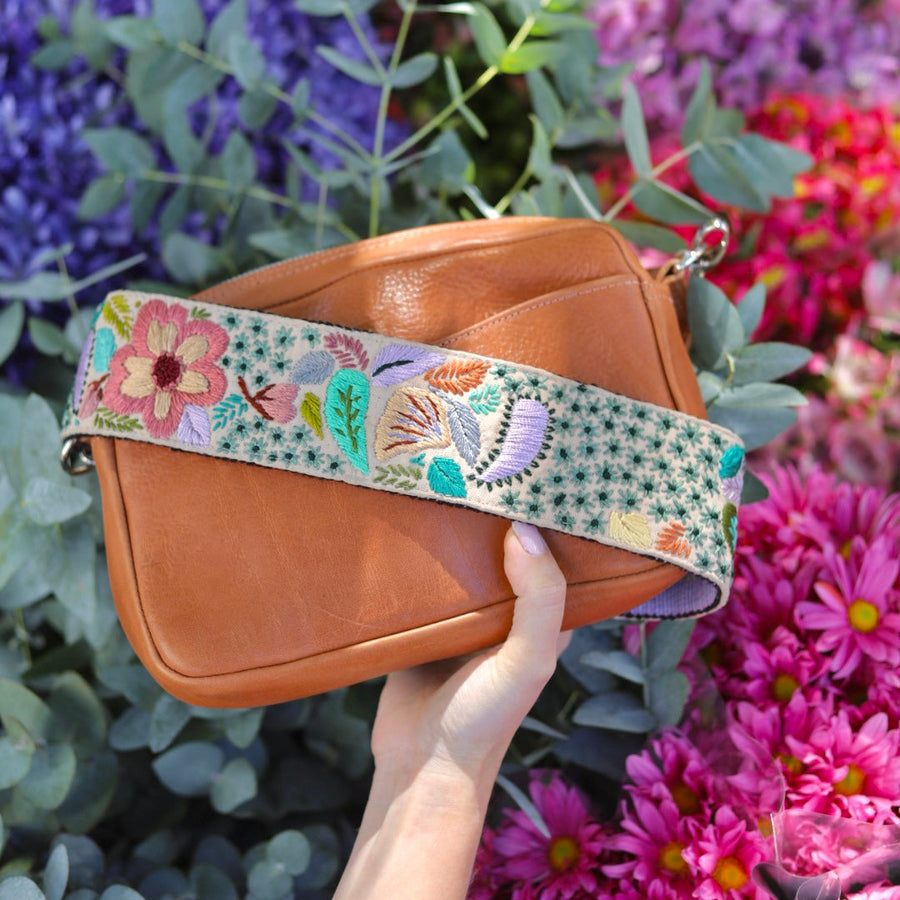 Brown leather handbag with a colorful floral strap held among flowers