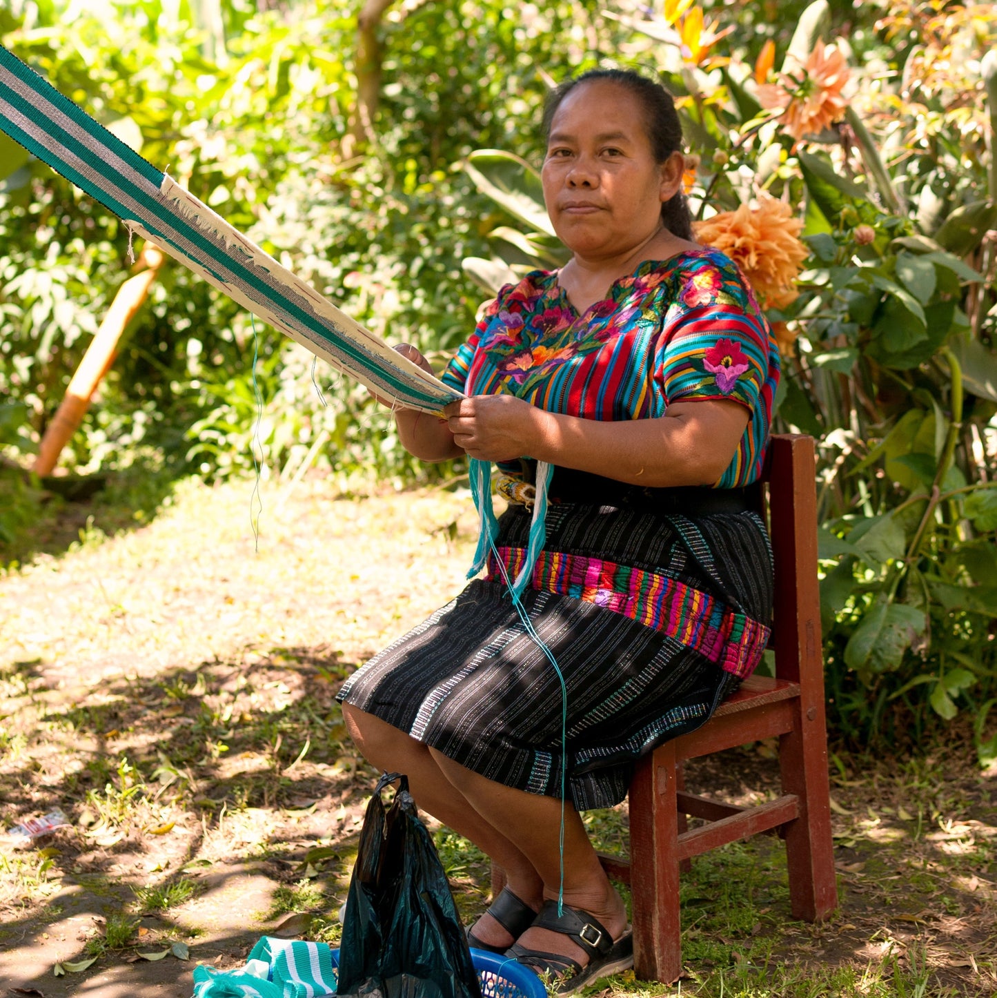 Woman sitting outdoors with a loom and yarn, surrounded by greenery.