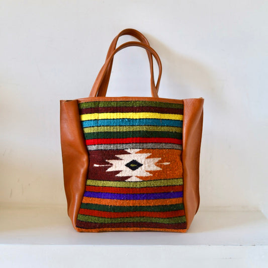 Tote bag with colorful striped pattern and brown leather sides on a white surface.